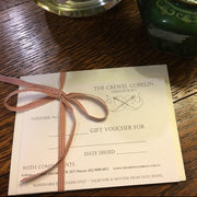 A gift voucher card from The Crewel Gobelin Needlecraft tied with a brown ribbon, placed on a wooden table next to a green ceramic vase with dried flowers.