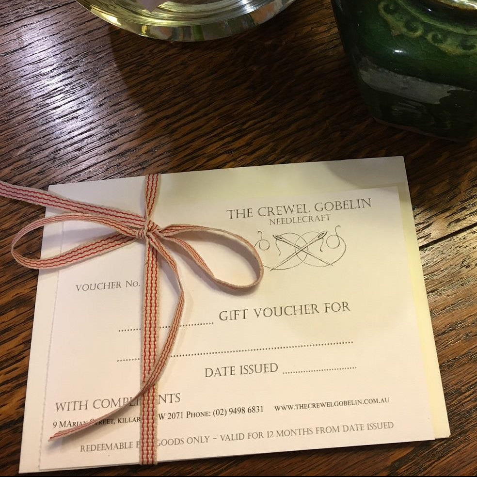 A gift voucher card from The Crewel Gobelin Needlecraft tied with a brown ribbon, placed on a wooden table next to a green ceramic vase with dried flowers.