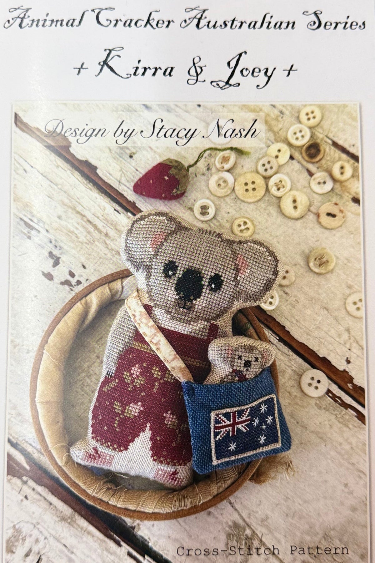 Cross-stitch pattern cover for 'Animal Tracker Australian Series + Kirra & Joey by Stacy Nash, featuring a finished cross-stitched koala wearing a red patterned sweater and a blue Australian flag pouch, surrounded by buttons and a strawberry on a rustic wood background.