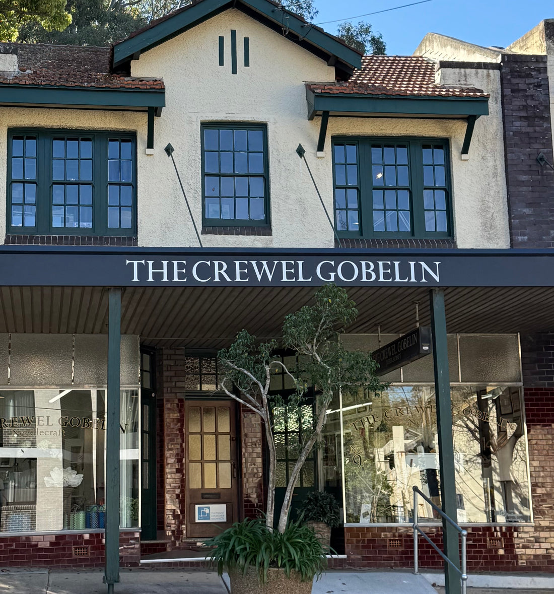 The Crewel Gobelin storefront with dark green awning, cream and brick facade, large windows, and potted plant
