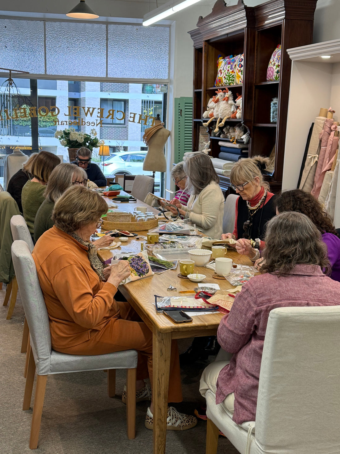 Group of women seated at wooden table doing needlework and craft projects in bright retail studio space