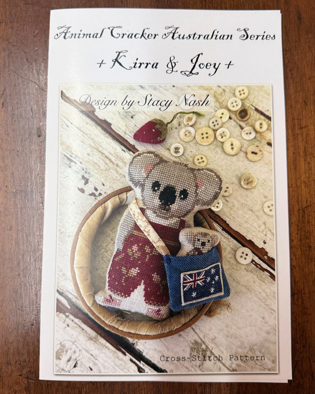 A cross-stitch pattern booklet titled 'Animal Cracker Australian Series + Kirra & Joey by Stacy Nash, featuring a finished cross-stitched kangaroo toy with a red-and-white striped body and a blue pouch, photographed on a wooden surface.