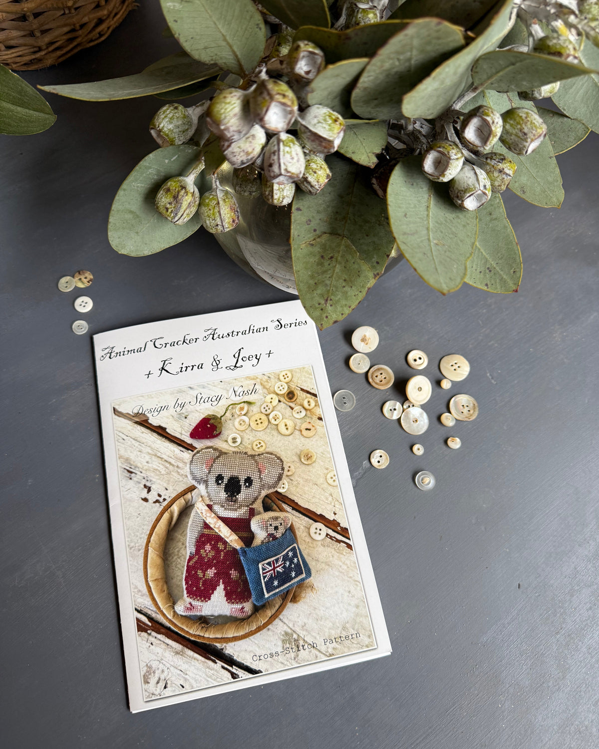 A flat-lay photograph of Animal Cracker Australian Series + Kira & Joey pattern booklet by Stacy Nash Designs, styled on a dark gray surface with decorative eucalyptus leaves and scattered buttons.